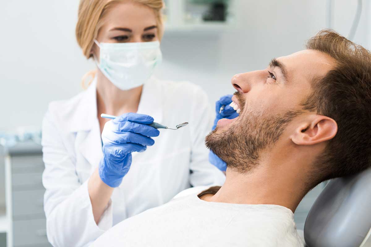 Young female dentist examining teeth of handsome smiling client in dental chair